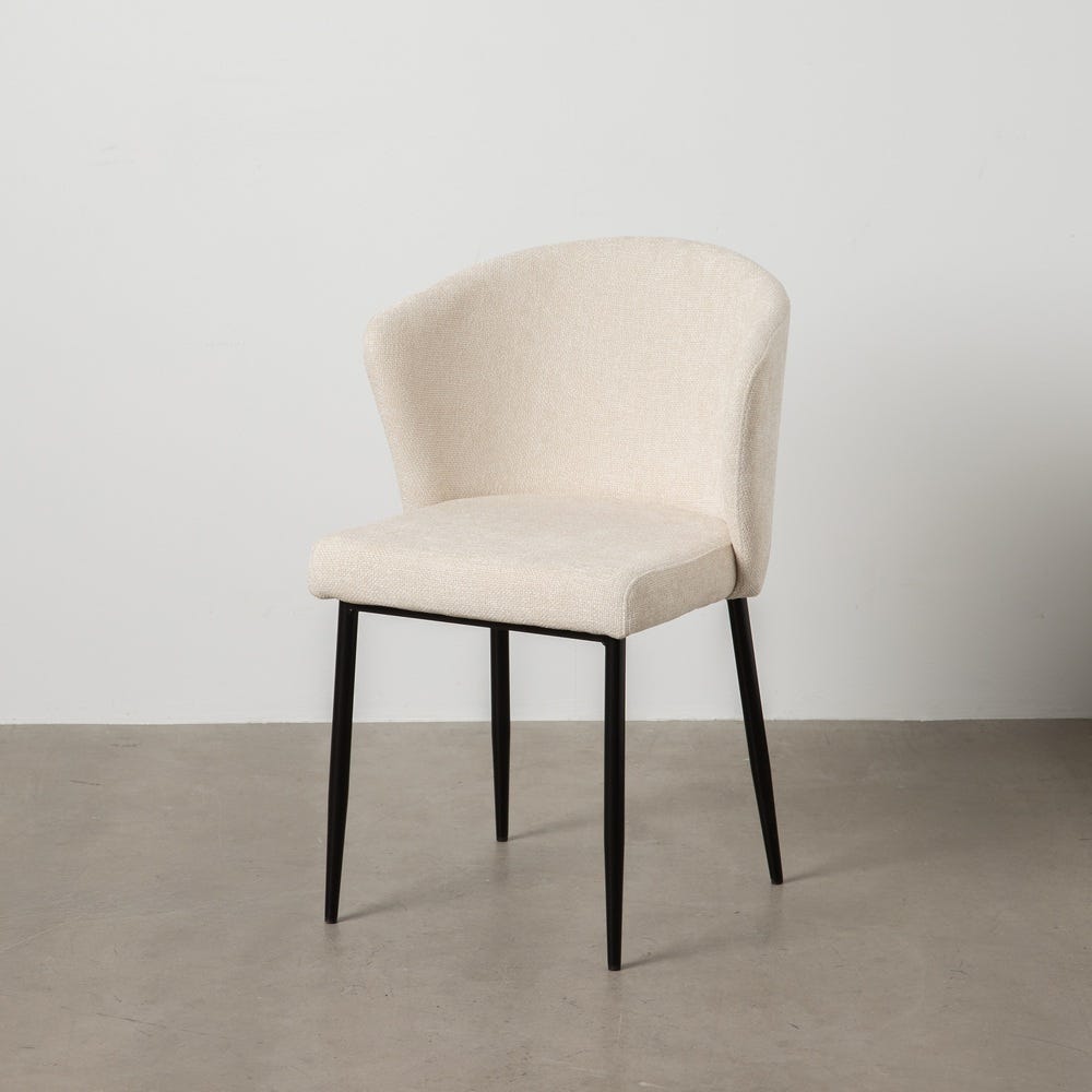 La "Silla de Comedor Tapizada Sev", con tapizado beige y patas de metal negro, irradia elegancia y comodidad mientras descansa graciosamente sobre un piso de concreto liso contra una pared blanca.
