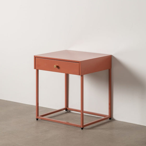 La Mesita de Noche Acero Industrial MET es una mesa auxiliar compacta y cuadrada en color rojo-naranja, que cuenta con un solo cajón y marco de metal cuadrado con diseño moderno, perfectamente colocada contra una pared blanca sobre un piso gris.