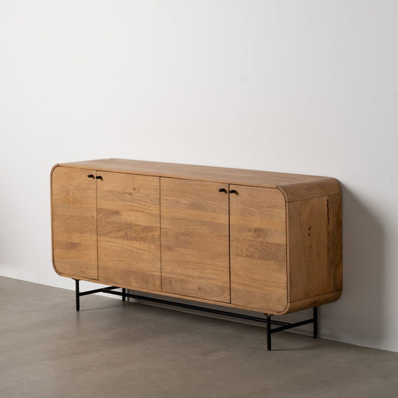 El Aparador Madera Mango Natural Mahón es un aparador moderno de madera de mediados de siglo con bordes redondeados, tres puertas tipo armario para almacenaje y un diseño moderno, con patas de metal negro. Se asienta elegantemente contra una pared blanca sobre un suelo de hormigón.