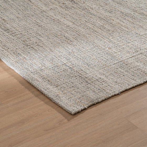 Primer plano de la esquina de una alfombra beige texturizada sobre un piso de madera, que presenta la delicada combinación de Lana Yute. La "Alfombra en Lana y Yute Nubes" agrega un estilo contemporáneo a cualquier espacio.