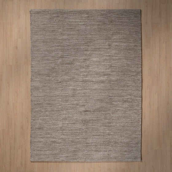 La "Alfombra en lana y yute Terraz", de diseño rústico y elegante, es una alfombra tejida rectangular de color gris con una sutil textura de rayas que reposa graciosamente sobre un suelo de madera clara.