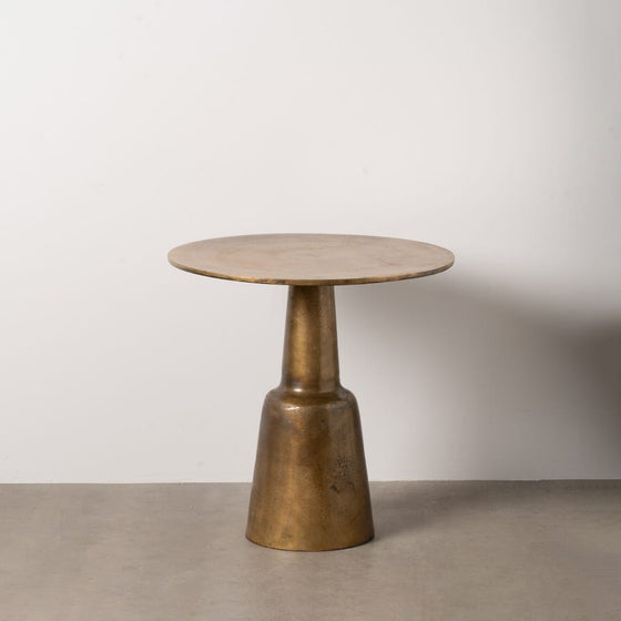 La Mesa auxiliar en Aluminio color cobre Alara Ø73, una pequeña mesa redonda metálica con base cónica, se alza sobre un suelo de hormigón contra una pared blanca lisa, mostrando su diseño moderno.