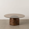 La Mesa de centro redonda (Ø80 cm) en madera de mango y cristal Miel presenta una tapa de cristal y una base cilíndrica de madera de mango, asentada sobre un suelo de hormigón contra una pared blanca, ejemplificando su diseño minimalista.
