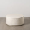 La Mesa de Centro en fibra de magnesio blanco 90x90 cm Fabro, redonda y moteada, reposa elegantemente sobre un suelo de hormigón contra una pared lisa.