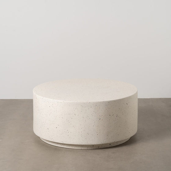 La Mesa de Centro en fibra de magnesio blanco 90x90 cm Fabro, redonda y moteada, reposa elegantemente sobre un suelo de hormigón contra una pared lisa.