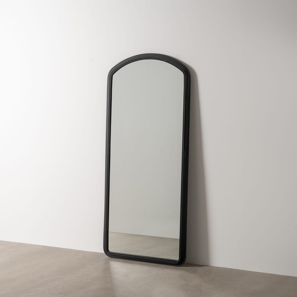 El Espejo Vestidor Metal-Cristal Negro Miras se apoya elegantemente sobre una pared blanca sobre el suelo de hormigón, destilando sofisticación.