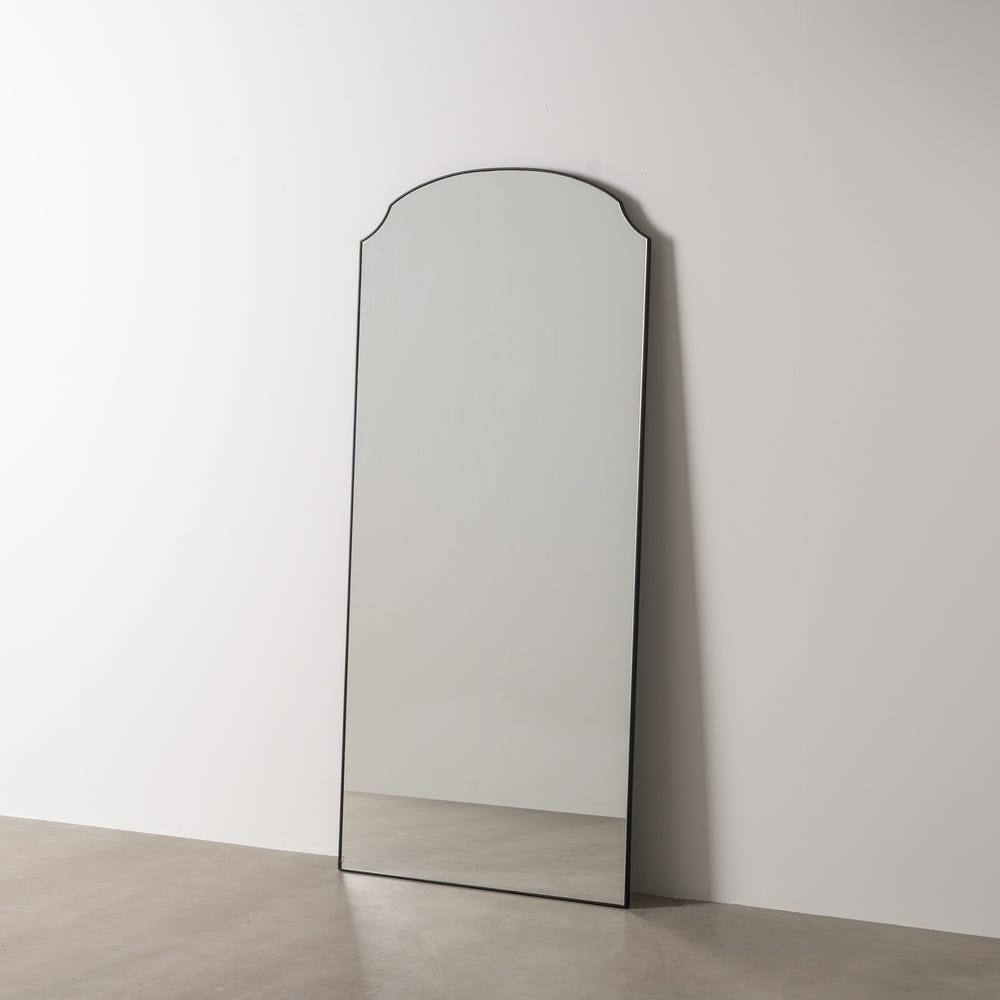 El alto Espejo Negro Metal-Cristal Vertical StyleX, con su forma rectangular y parte superior redondeada, se apoya contra una pared blanca lisa sobre un piso desnudo, mostrando sofisticación moderna en su diseño minimalista.