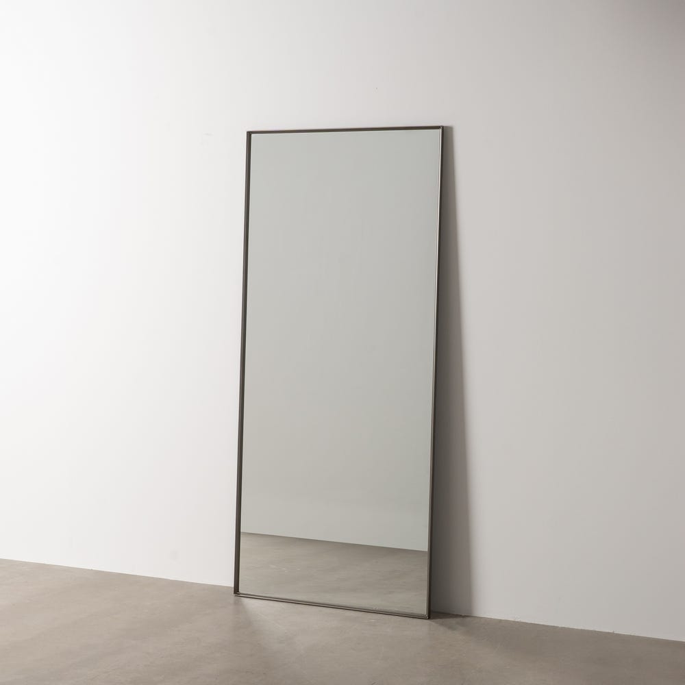 El Espejo Acero Metal-Cristal Walli, un espejo alto y rectangular con un elegante marco negro, se apoya contra una pared blanca sobre un piso de concreto, ejemplificando el diseño moderno.