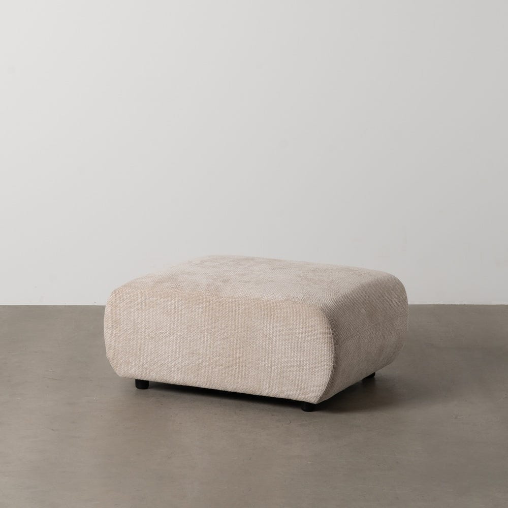 Un Puf Cuadrado en Tejido beige Aria rectangular de color beige con esquinas redondeadas descansa sobre un piso de concreto desnudo contra una pared blanca lisa, capturando el diseño contemporáneo al mismo tiempo que brinda comodidad y estilo.