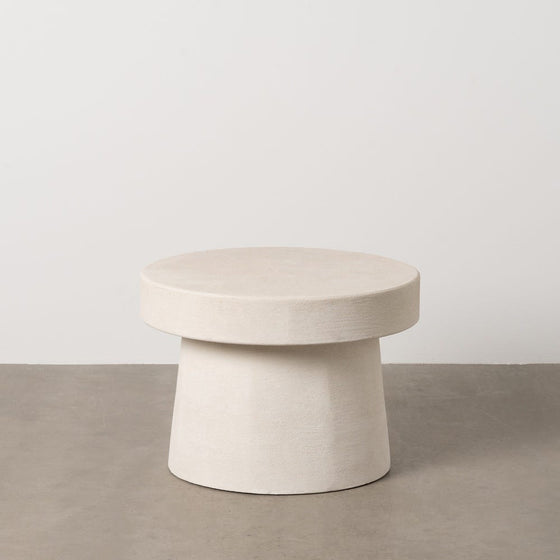 La Mesa Auxiliar Cilíndrica en DM Blanco Wilma Ø58, de diseño minimalista y base cónica, reposa sobre el suelo de hormigón. Su estilo moderno realza la decoración minimalista sobre una pared blanca.
