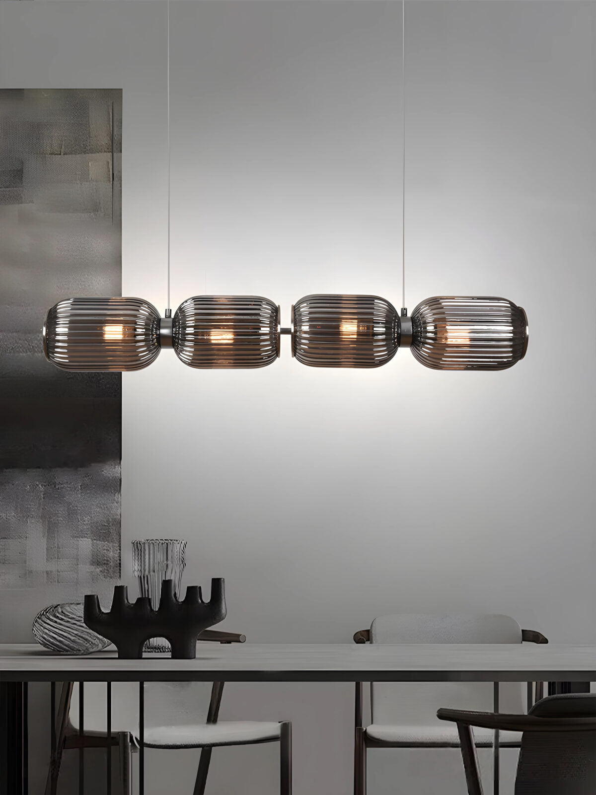 Un comedor moderno cuenta con una mesa de cristal, sillas y un centro de mesa escultural. Sobre la mesa cuelga la lámpara Kairo 4 de techo en acero y vidrio con múltiples focos, que proporciona una iluminación uniforme en su diseño cilíndrico contemporáneo.