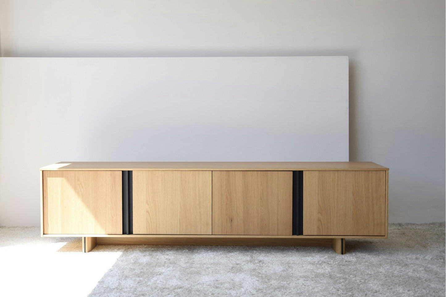 El mueble TV en DM y roble detalles en metal negro Clivo presenta un diseño contemporáneo con cuatro puertas y elegantes tiradores negros, elegantemente colocados contra una pared blanca lisa sobre una alfombra clara.