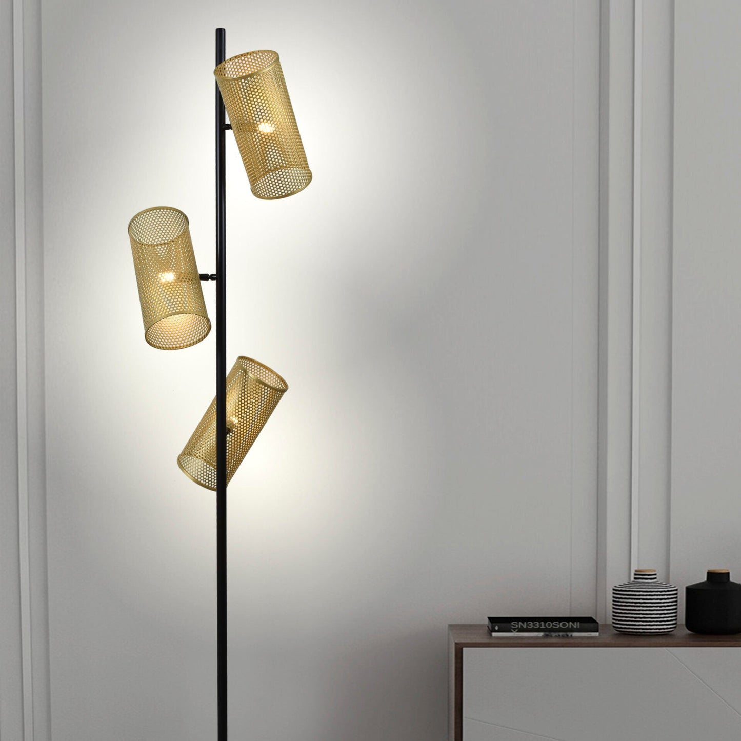 La Lámpara de pie de acero Grid 3, con tres pantallas de malla cilíndricas y un estilo industrial, se encuentra sobre un suelo de baldosas junto a un mueble blanco minimalista.