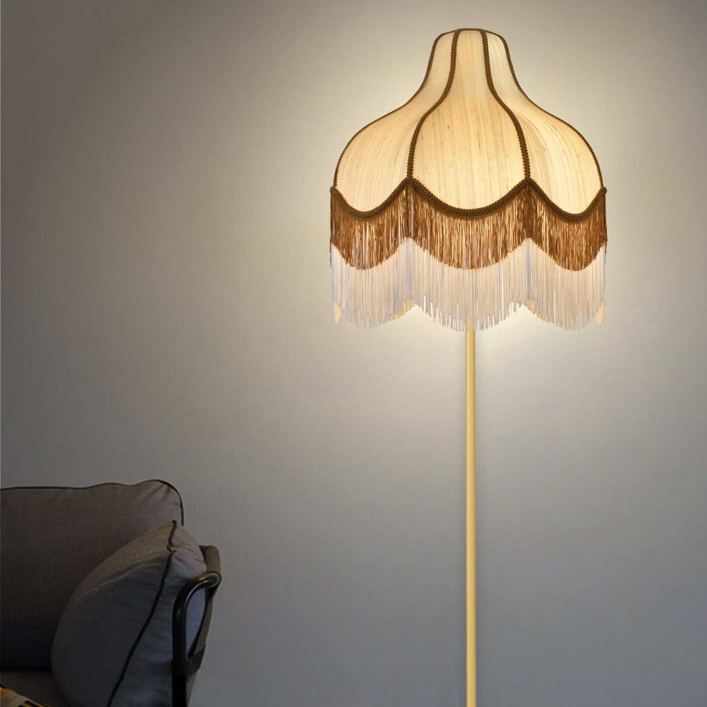 La Lámpara de pie vintage de flecos Bez con pantalla beige con volantes ilumina una habitación tenue. Un sofá gris es parcialmente visible a la izquierda, resaltando el diseño elegante del espacio.
