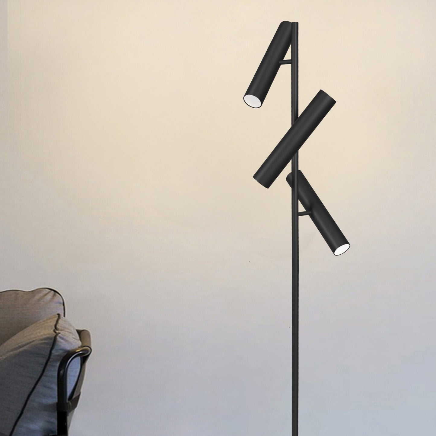 La Lámpara de pie de acero Toru 3 en color negro ofrece una elegancia industrial con sus tres luces cilíndricas orientables, complementando perfectamente un sillón gris contra una pared de color claro.