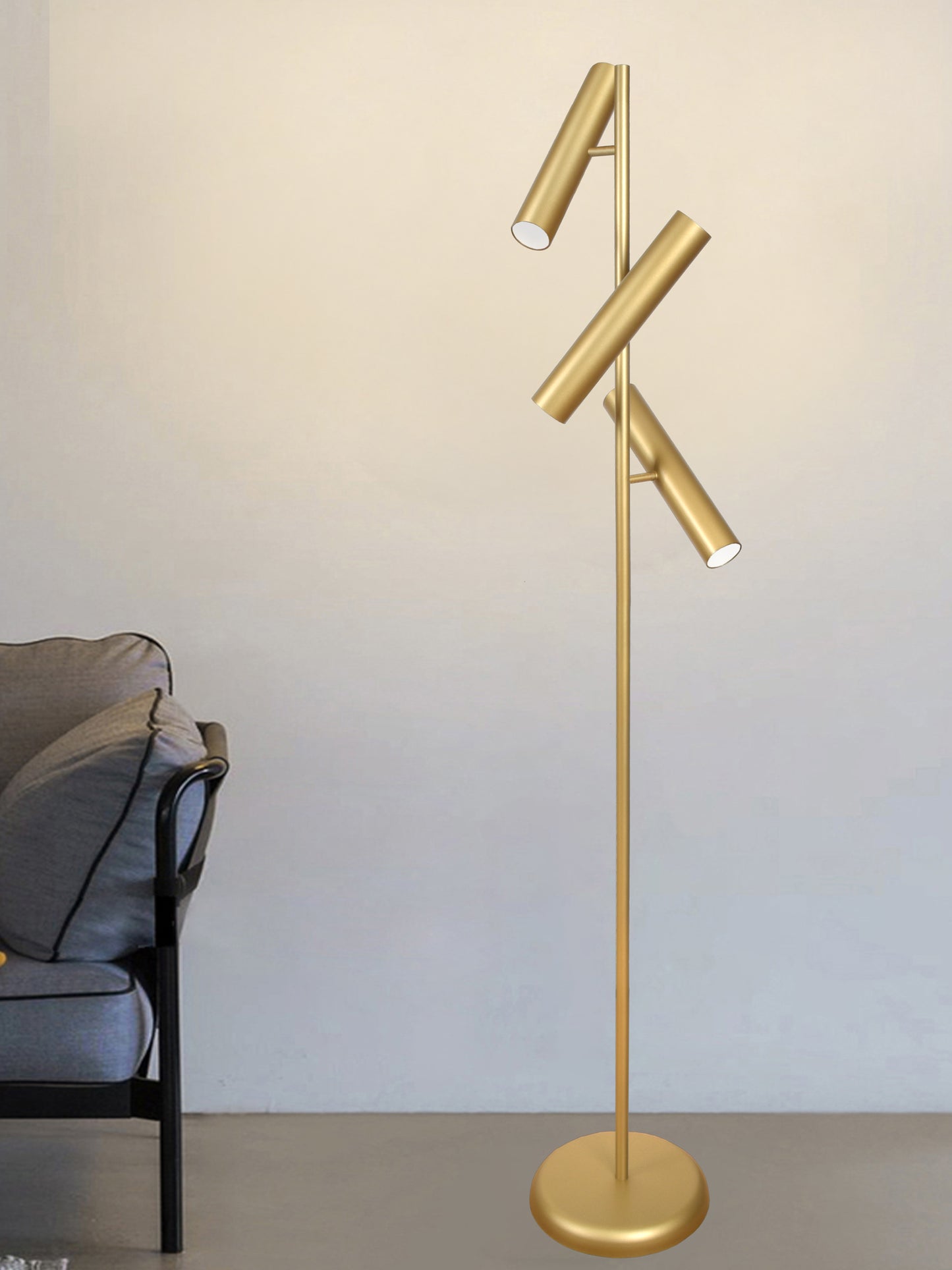 La lámpara de pie de acero Toru 3, con un moderno diseño dorado con tres luminarias cilíndricas ajustables, se encuentra junto a una silla acolchada gris contra una pared sencilla y rezuma elegancia industrial.