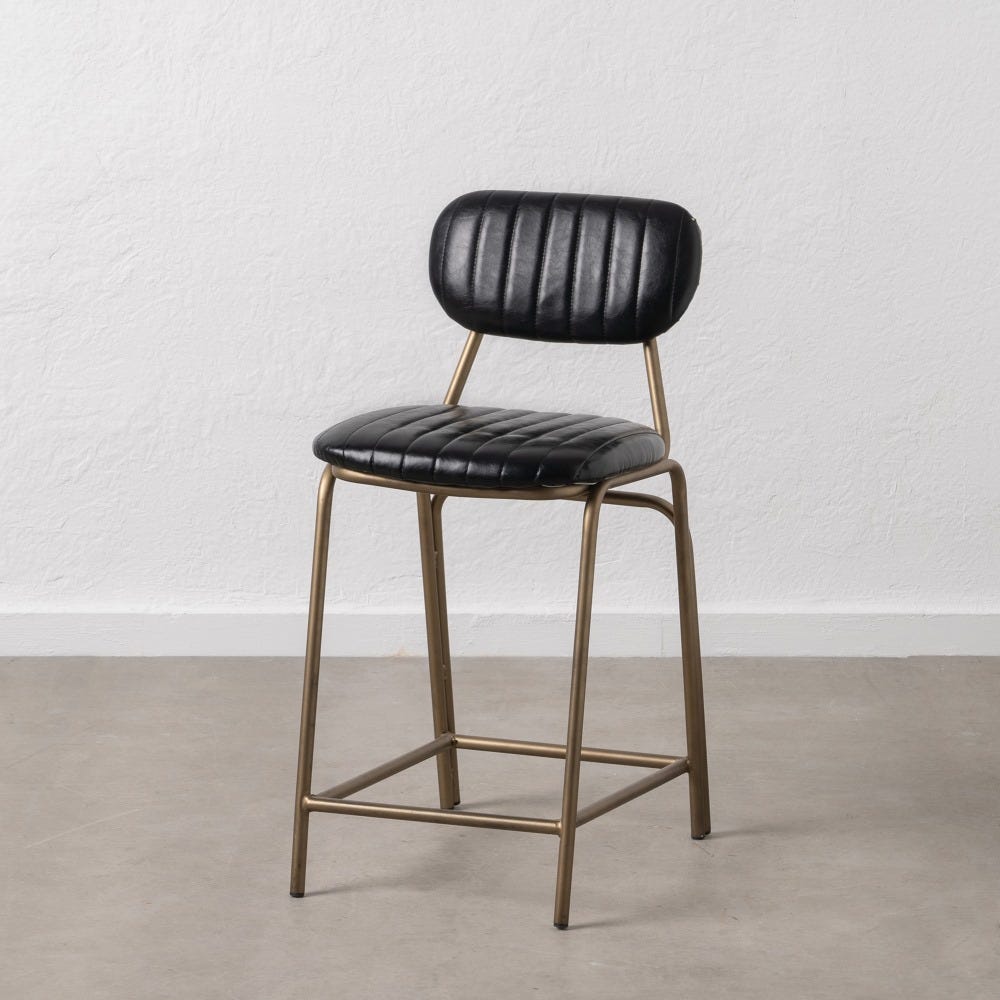 El Taburete alto (88 cm) en cuero sintético y metal Magnus, con asiento y respaldo acanalados en negro y dorado, se alza elegantemente sobre un piso gris contra una pared blanca.