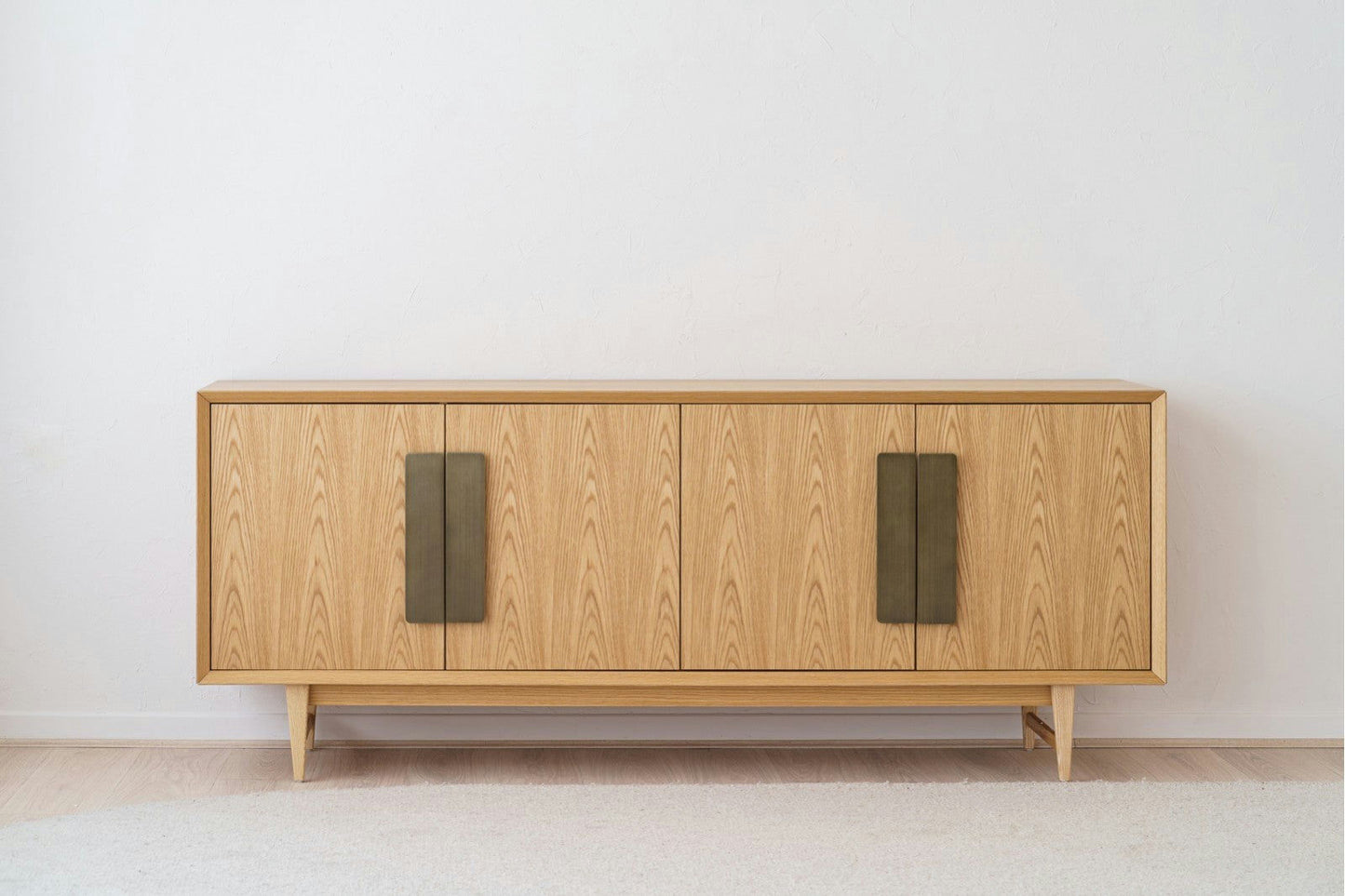 El Aparador de Roble y Latón OIA, elaborado en elegante madera de roble, cuenta con cuatro puertas verticales con tiradores envejecidos de latón oscuro que realzan bellamente una pared blanca lisa.