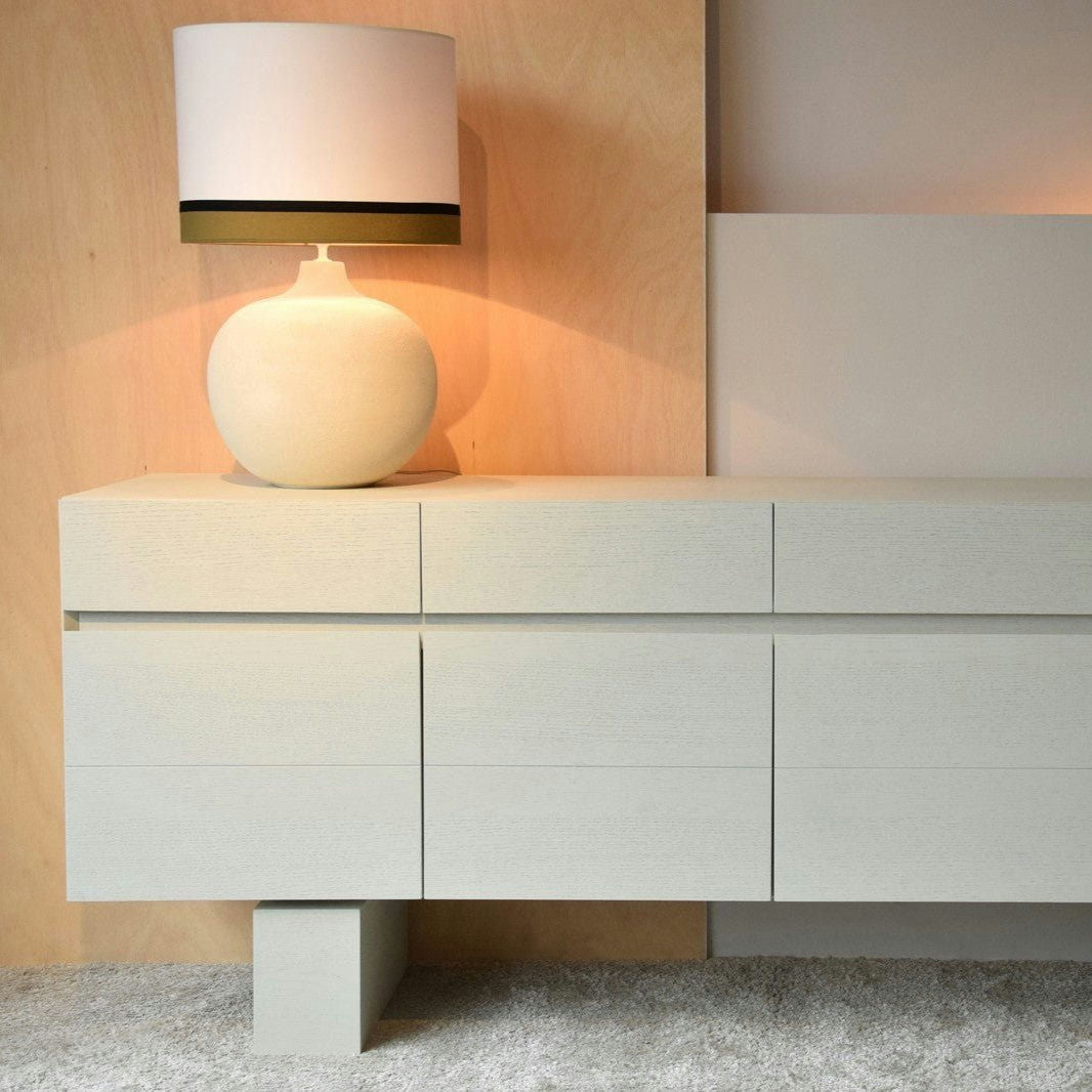 El Aparador Moderno en Madera Blanco Roto Alba es una pieza contemporánea de líneas limpias y generoso espacio de almacenaje en sus cajones. Presenta un acabado en color claro, complementado por una sencilla lámpara blanca con base redonda y pantalla cilíndrica en la parte superior.
