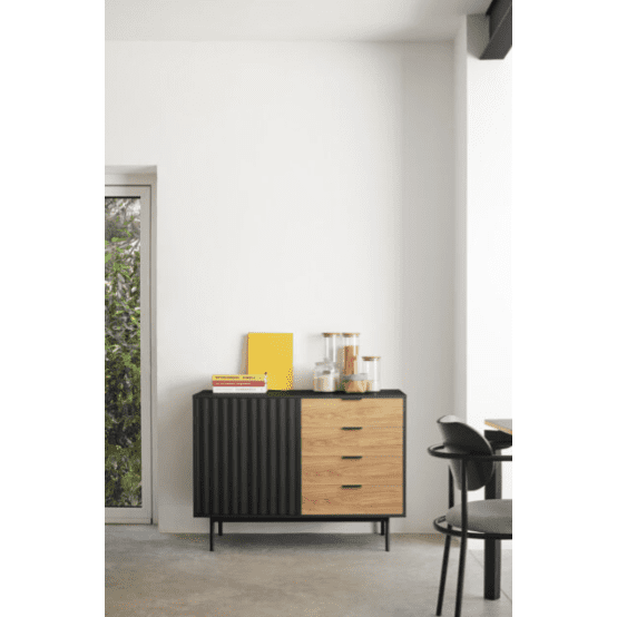 El Aparador Sierra 1P4C con diseño en negro y madera, adornado con libros y frascos, ubicado en una habitación minimalista al lado de una puerta abierta, muestra un ejemplo de diseño de interiores.