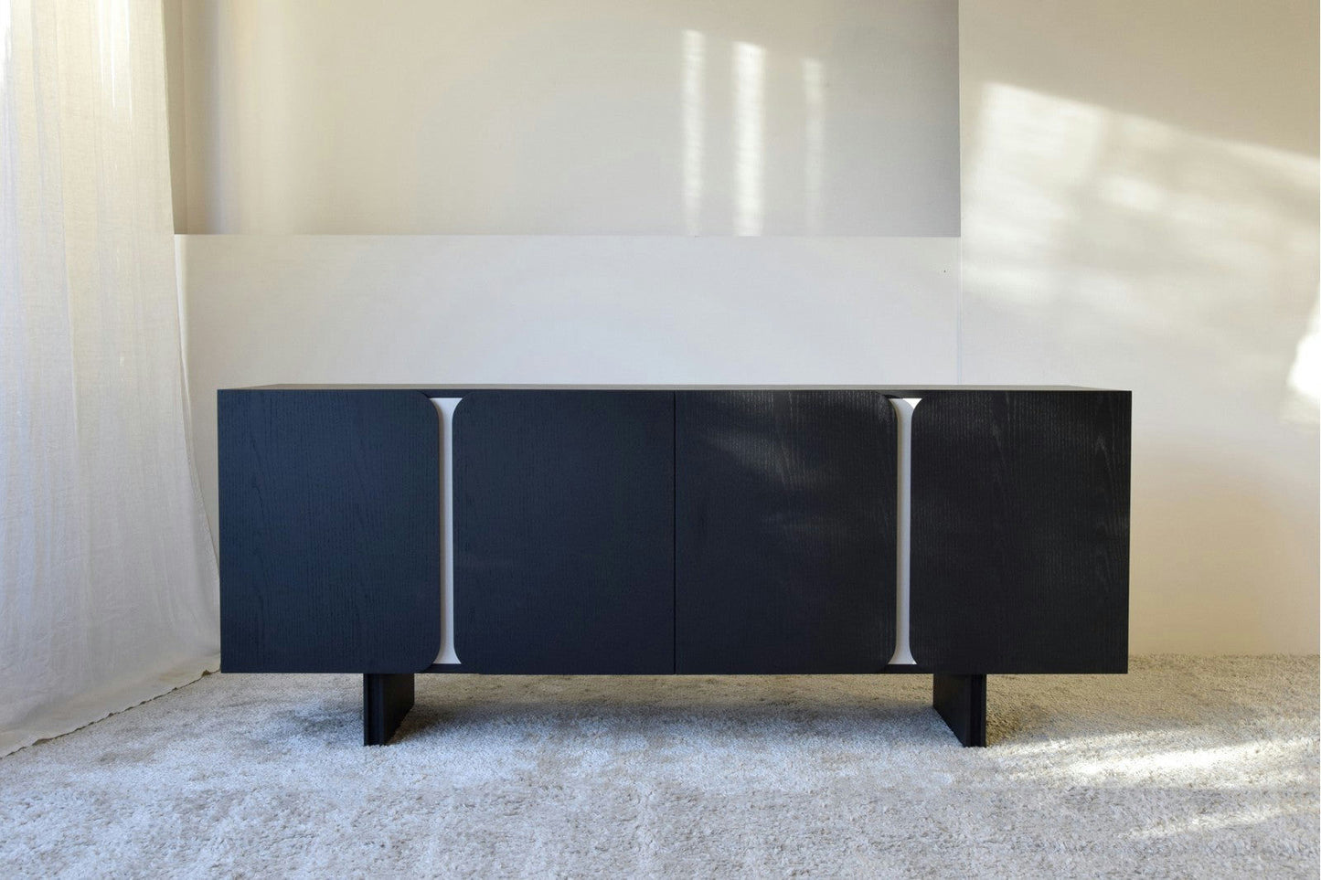 El Aparador bajo en Madera Negro Mate y Arena Curva de la Colección Curva luce un moderno acabado en negro con tres paneles verticales, que se alzan elegantemente sobre una alfombra clara en una habitación luminosa. Su diseño contemporáneo complementa perfectamente cualquier interior elegante.