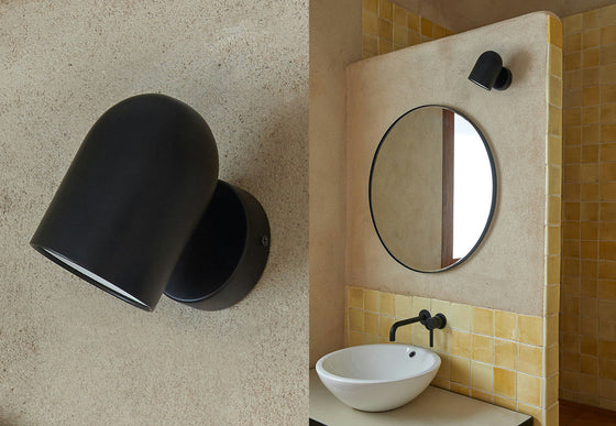 Una elegante lámpara de pared Pipe de color negro mate se encuentra junto a un baño con un espejo redondo, un lavabo blanco y un grifo negro sobre azulejos beige y amarillos. Esta iluminación funcional añade un toque moderno al elegante espacio.