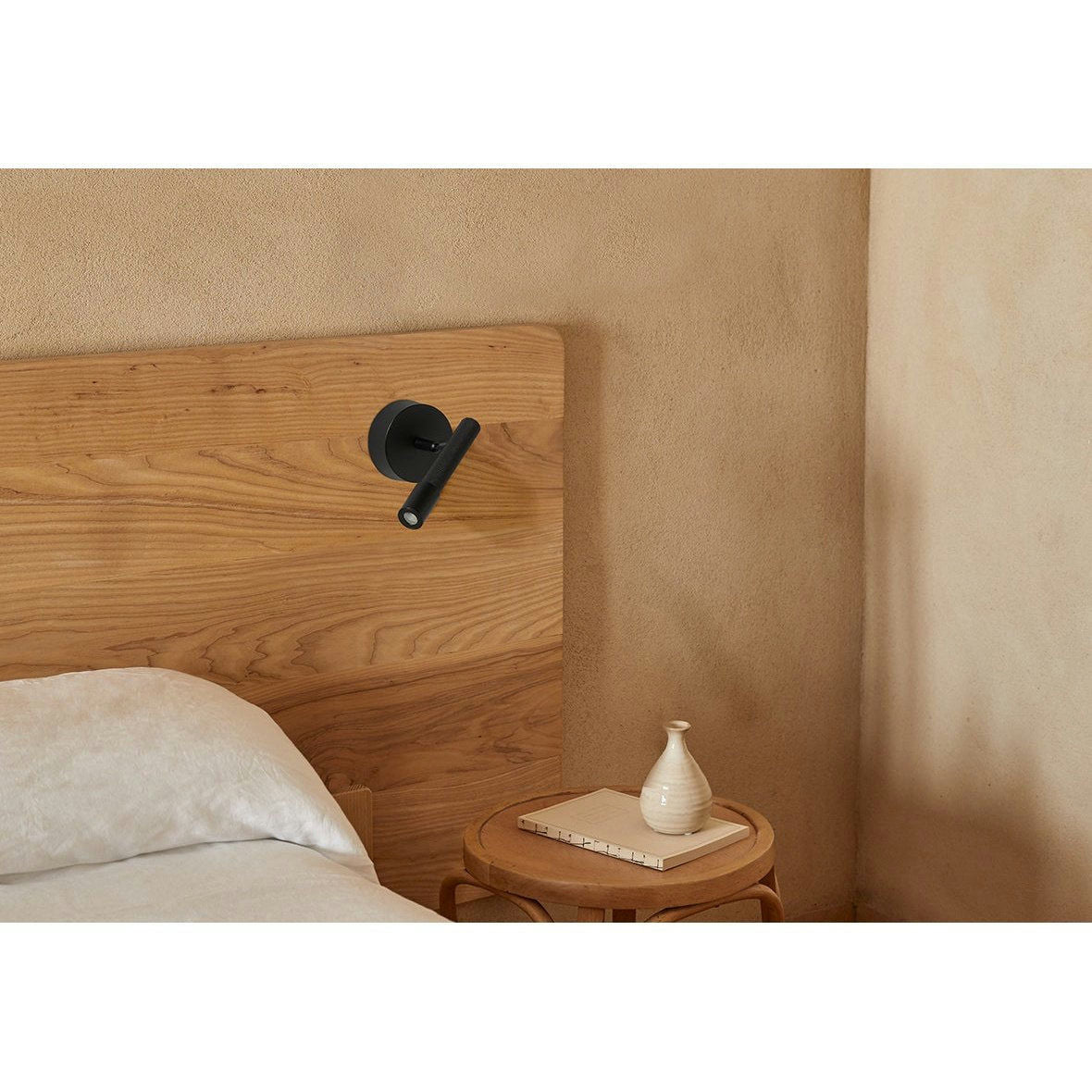Un dormitorio minimalista cuenta con un cabecero de madera y una mesa auxiliar. Sobre la mesa reposa un jarrón blanco, mientras que sobre la cama se encuentra la lámpara de pared moderna Ycro de color negro con tecnología LED.