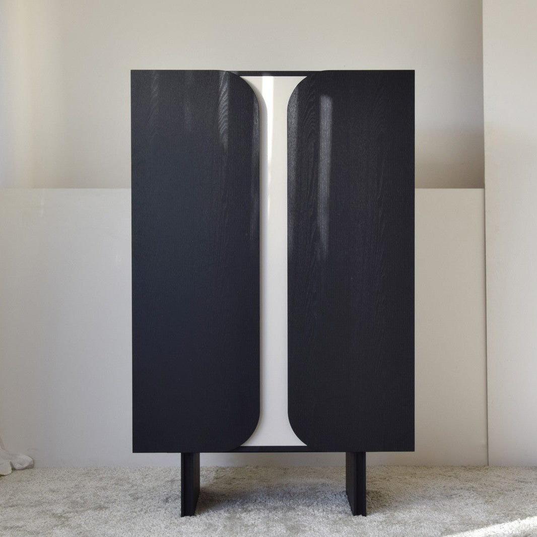 El Armario Roble Negro Mate y Detalle Arena de la Colección Curva presenta un elegante gabinete negro con puertas curvas, que incorpora un diseño contemporáneo minimalista mientras se coloca elegantemente sobre una alfombra clara en una habitación sencilla y de tonos neutros.