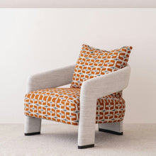  La butaca tapizada en beige y naranja doble presenta un diseño moderno con cojines con estampados geométricos en naranja y blanco, apoyabrazos y patas en gris claro, confeccionados en 100 % poliéster. Destaca hermosamente contra una pared blanca lisa y un piso alfombrado.