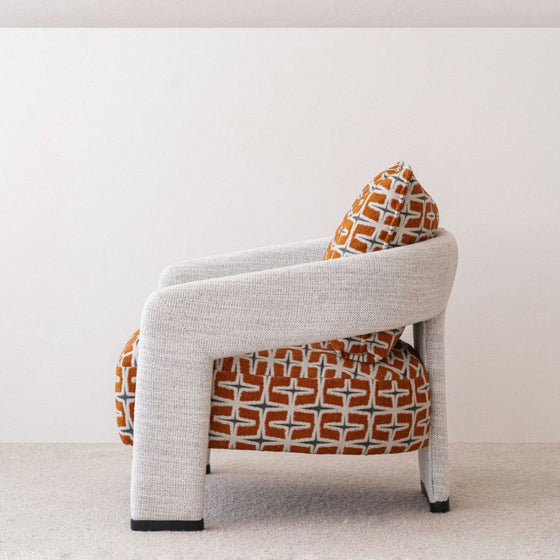 Colocada sobre una alfombra beige, la Butaca Tapizada en Beige y Naranja Doble tiene un patrón geométrico moderno con tela 100% poliéster en tonos naranja y gris, acolchado grueso y apoyabrazos curvos.