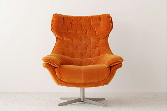 La Butaca Giratoria Tapizada en Terciopelo Orange presenta un diseño moderno con una elegante base giratoria de metal, elegantemente situada sobre un piso alfombrado claro contra una pared blanca lisa, personificando el mueble elegante.