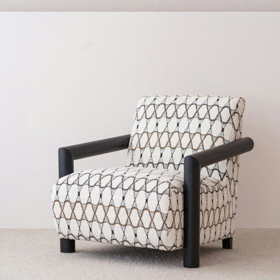 La Butaca en madera de Roble y Bouclé Gema, que presenta un patrón geométrico de diseños de diamantes en blanco, negro y marrón, está colocada contra una pared lisa con apoyabrazos cilíndricos negros.
