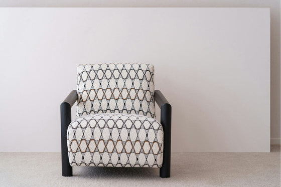 La Butaca en madera de Roble y Bouclé Gema, con un patrón geométrico y brazos de roble macizo oscuro, se asienta elegantemente sobre una alfombra clara contra una pared lisa de color claro.
