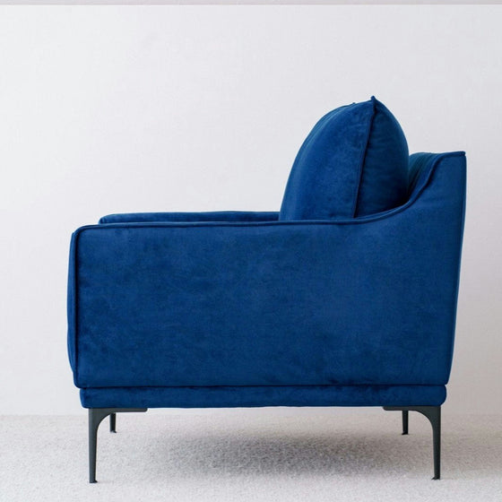 La Butaca en Terciopelo Azul Noche Helia descansa elegantemente contra una pared clara sobre una alfombra texturizada, personificando el lujo y la comodidad que eleva cualquier estilo de interiorismo.