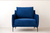 La butaca en terciopelo azul noche Helia, con cojines mullidos y patas de metal negro, aporta lujo y comodidad al interiorismo frente a la pared blanca.