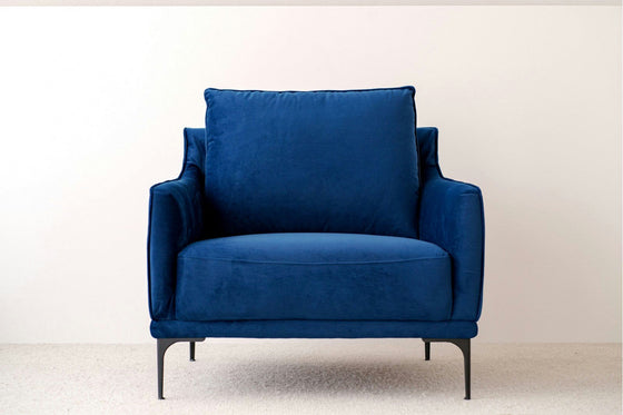 La butaca en terciopelo azul noche Helia, con cojines mullidos y patas de metal negro, aporta lujo y comodidad al interiorismo frente a la pared blanca.