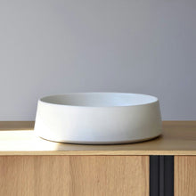  El Centro de Mesa Minimalista Blanco Mate Cerámico, un recipiente de cerámica blanca con acabado mate, se destaca como pieza central en un gabinete de madera clara contra una pared gris simple, incorporando un diseño minimalista.