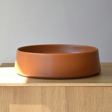  El Centro de Mesa Minimalista Ocre Mate Cerámico, un recipiente de cerámica redondo y plano, sirve como una elegante pieza central sobre una superficie de madera sobre un fondo liso.