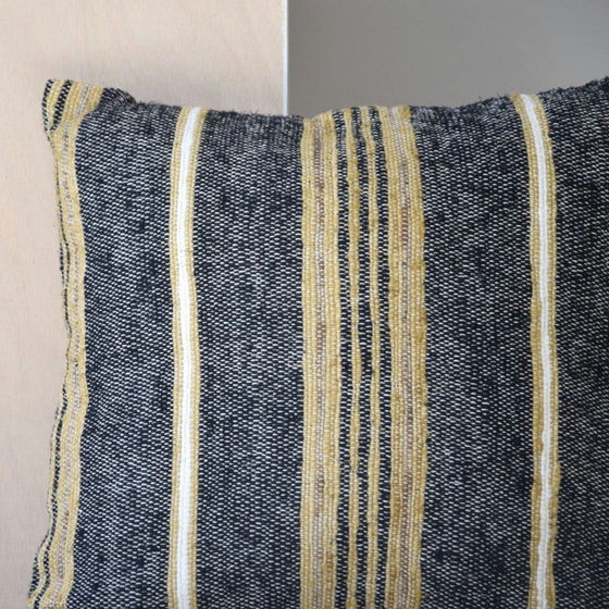El Cojín Adra N.3 Algodón Negro y Camel, elaborado 100% algodón, presenta un diseño texturizado de rayas verticales doradas y negras que complementa maravillosamente las paredes de colores claros.
