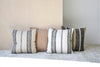 Cuatro cojines Cojín Adra N.3 Algodón Negro y Camel se presentan con estilo sobre una alfombra clara contra una pared lisa. Fabricados en 100 % algodón, realzan la elegancia del diseño de la habitación con sus tonos neutros de rayas.