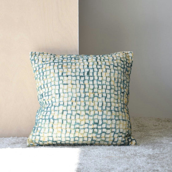 Un Cojín Algodón Verde Blanco de la colección Vega N.2, con un patrón tejido en tonos azules y amarillos, reposa sobre un panel de madera sobre un suelo alfombrado, realzando la decoración de interiores.