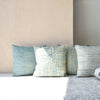 El Cojín Algodón Verde Blanco. Vega N.2, un conjunto de cuatro almohadones texturizados en tonos azul y crema, está bellamente dispuesto sobre una alfombra clara contra una pared lisa, ideal para la decoración de interiores.