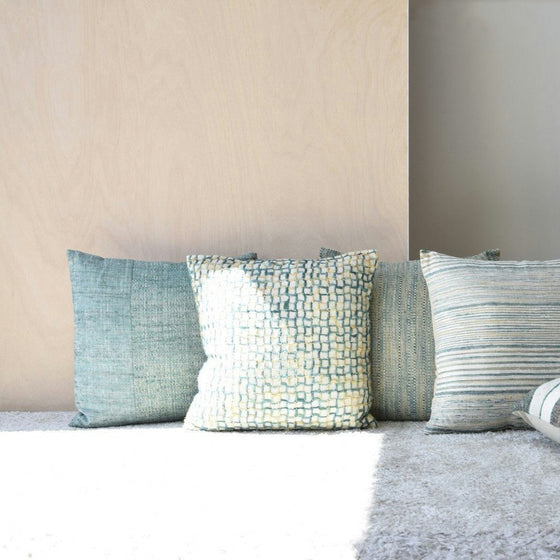 El Cojín Algodón Verde Blanco. Vega N.2, un conjunto de cuatro almohadones texturizados en tonos azul y crema, está bellamente dispuesto sobre una alfombra clara contra una pared lisa, ideal para la decoración de interiores.