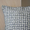 El Cojín cuadrado de Algodón Verde-Gris OZA N.3, elaborado en algodón 100% de alta calidad, presenta un patrón de cuadrícula texturizada en tonos blancos y grises, realzando perfectamente el fondo de madera y beige para una decoración sofisticada.