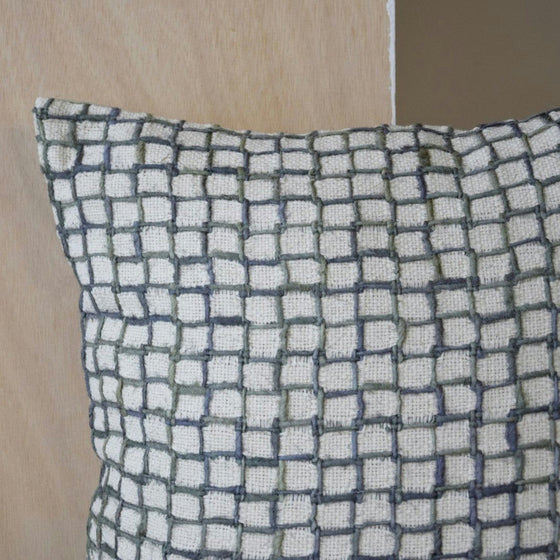 El Cojín cuadrado de Algodón Verde-Gris OZA N.3, elaborado en algodón 100% de alta calidad, presenta un patrón de cuadrícula texturizada en tonos blancos y grises, realzando perfectamente el fondo de madera y beige para una decoración sofisticada.