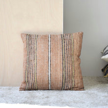  El cojín Adra N.4 - Algodón Tonos Ocre y Mostaza de la exclusiva colección Adra presenta rayas naranjas, marrones y beige. Reposa elegantemente sobre una alfombra clara contra un panel de madera, agregando un toque acogedor a cualquier habitación.