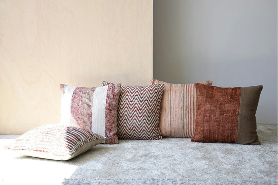Cinco almohadas estampadas en tonos neutros, incluido el elegante Cojín Mara N.4 en Algodón Tostado y Rojo, descansan sobre una alfombra gris claro contra una pared lisa, mostrando un sofisticado diseño de interiores.