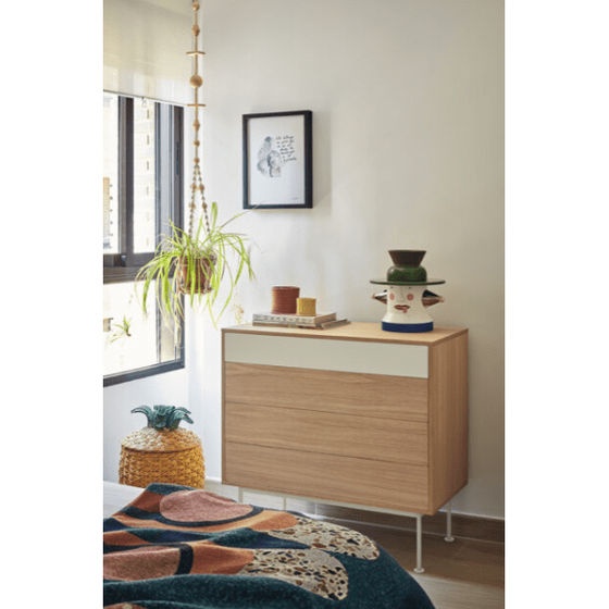 Una pequeña Cómoda en MDF y Roble con 4 Cajones Yoko 4C se encuentra debajo de la ventana, personificando el diseño minimalista. Está adornada con una planta en maceta, arte enmarcado, una escultura de cerámica y una canasta de piña en la esquina del dormitorio.