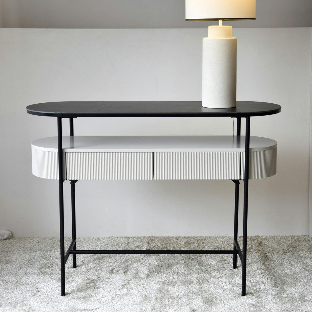 La Consola Gris arena con Piedra Sinterizada y Metal Negro Urbana N.24 marca tendencia en interiorismo, presenta una consola moderna con una elegante tapa ovalada negra y un cajón blanco acanalado, sostenido por delgadas patas negras, y personifica el diseño contemporáneo.