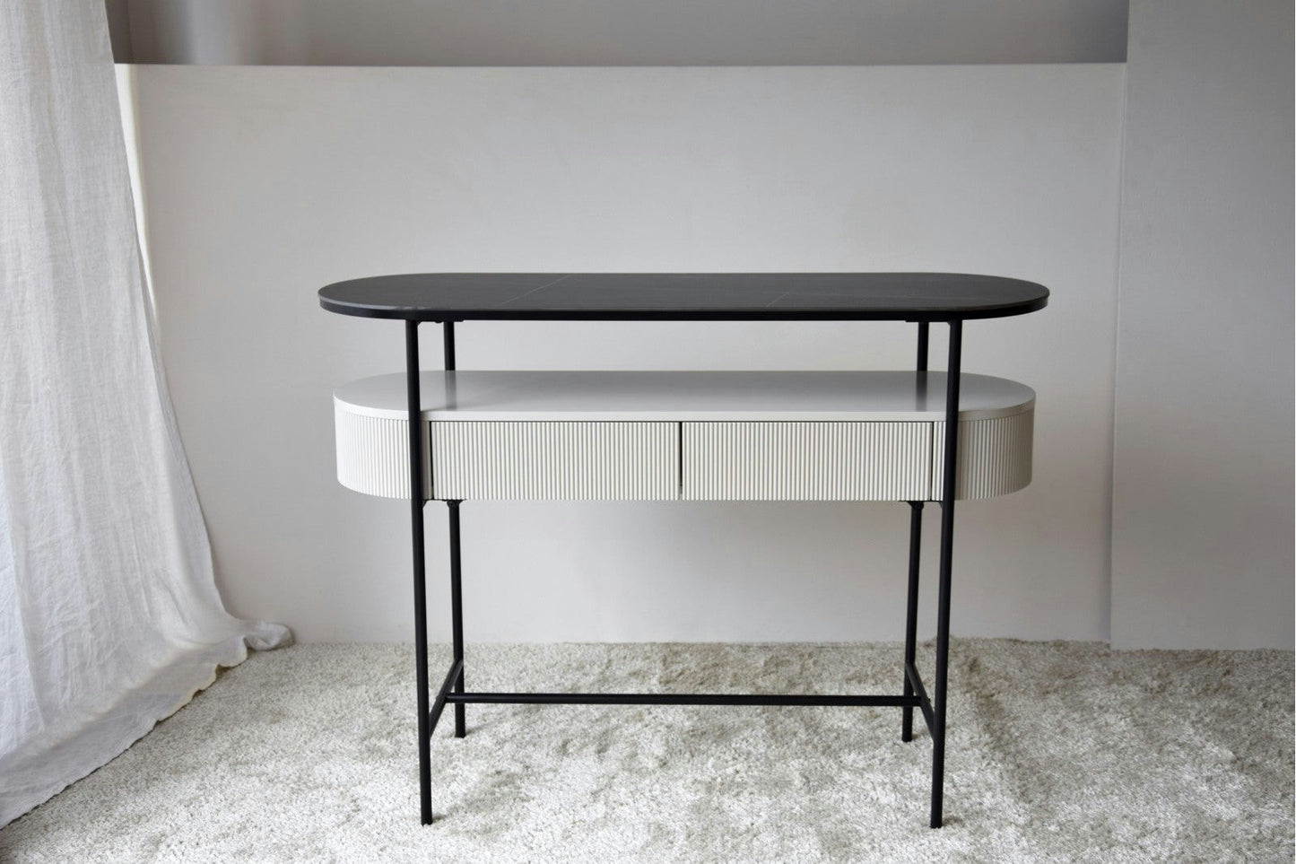 La Consola Gris arena con Piedra Sinterizada Urbana N.24 es una consola contemporánea con un elegante tablero de piedra sinterizada gris y patas de metal negro, que armoniza con las tendencias del interiorismo en diseño contemporáneo, elegantemente colocada sobre un piso alfombrado claro.